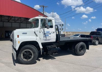 A large L8000 flat bed service truck custom painted white with our MCIS logo decal placed on the door. The hydraulic blat bed is coated in high quality bedliner protective coating by Vortex Sprayliners