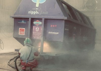 A blast media specialist spraying a large trash dumpster