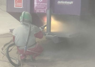 A blast media specialist spraying a large trash dumpster